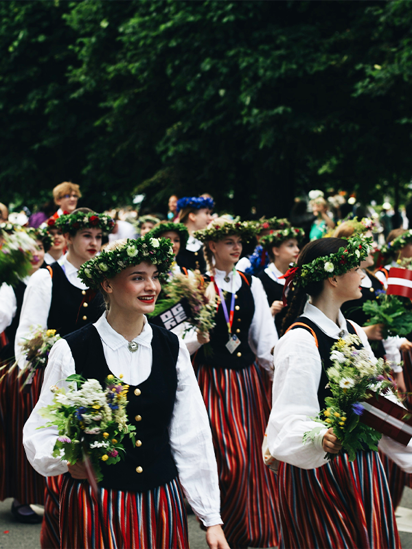 Young women in traditional Latvian folk costumes with floral wreaths and wildflower bouquets smiling during a street parade in Riga