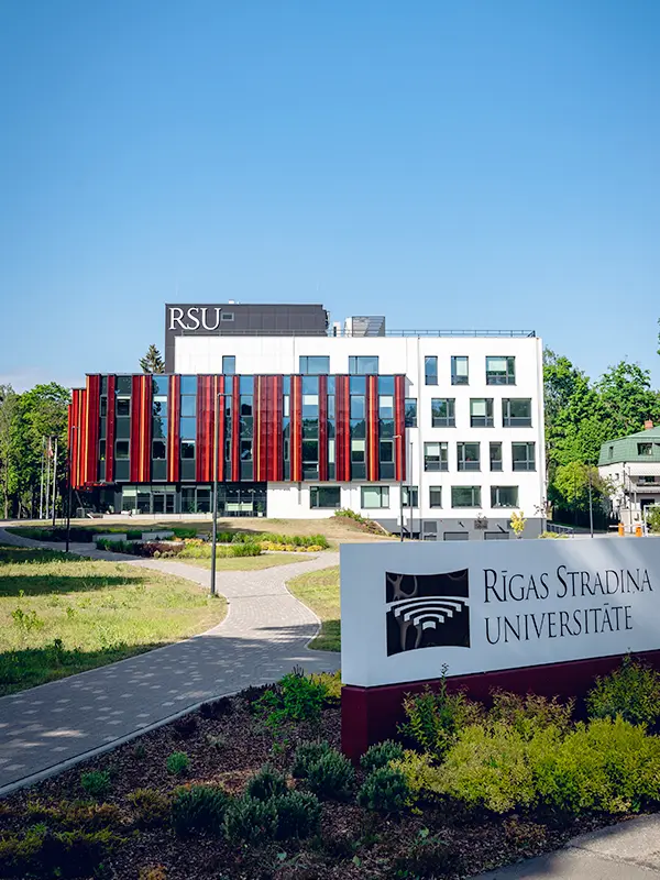 Modern white RSU building with red vertical panels and Rīgas Stradiņa Universitāte sign surrounded by green gardens on a sunny day