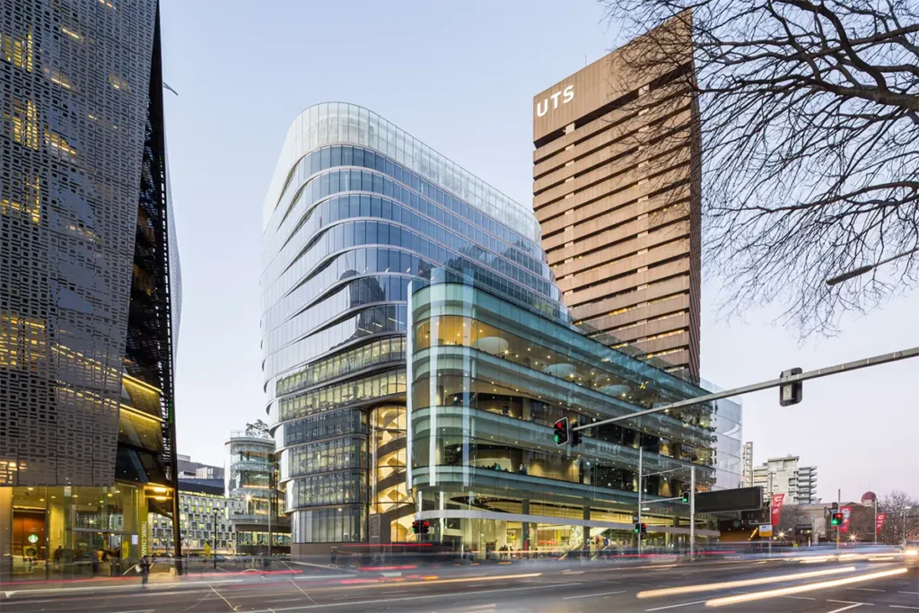 Curved glass UTS buildings glowing at dusk along a busy Sydney CBD intersection with light trails from passing traffic