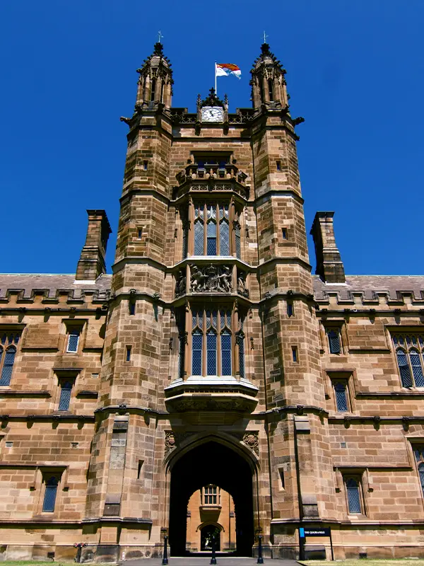 Close-up view of the Gothic sandstone gateway tower at the University of Sydney Quadrangle, with twin spires, a clock, and the university flag flying above the arched entrance; a top destination for international students studying abroad in Australia.