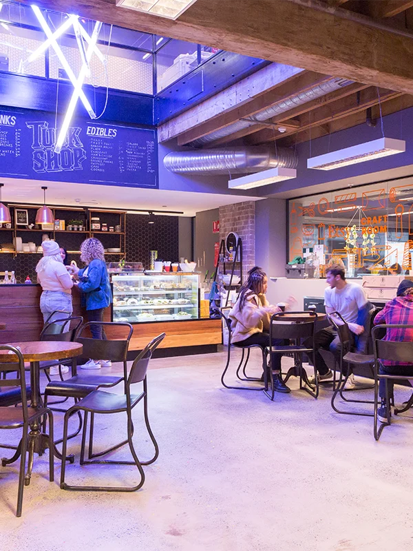 Students sitting at industrial style tables inside the Tuck Shop café at Torrens University Adelaide with blue neon lighting and exposed wooden beams overhead